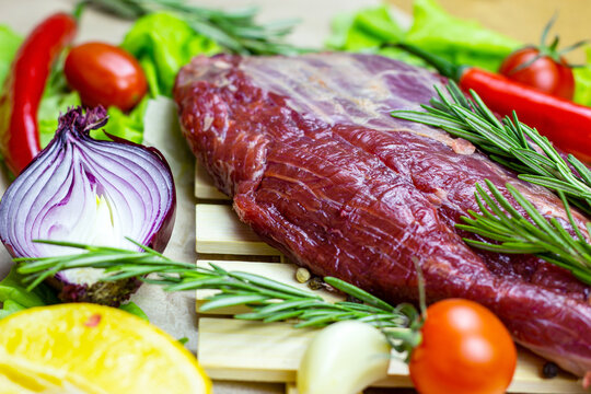 Raw red beef meat with green rosemary and fresh vegetables on light wooden cutting board background.