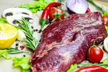 Raw red beef meat with green rosemary and fresh vegetables on light wooden cutting board background.