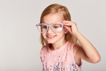 Portrait 4-5 year old kid girl posing in pink glasses and dress over grey background with copy space