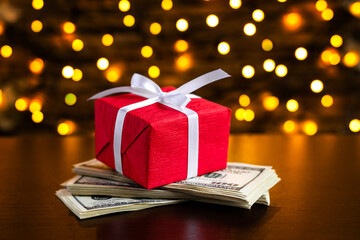 Red gift box on the USA 100 dollar bills on blurred lights background. American dollar. Christmas and New Year money present. Gift cost. Hundred. Finance and business concept. Christmas shopping