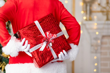 Santa Claus is holding a gift in a red package behind his back