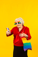 Studio portrait of a senior woman wearing a red shirt, rainbow sunglasses and a bag, having a video call with her mobile phone, against a yellow background