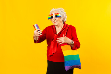 Fototapeta na wymiar Studio portrait of a senior woman wearing a red shirt, rainbow sunglasses and a bag, having a video call with her mobile phone, against a yellow background