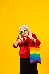 Studio portrait of a senior woman wearing a red shirt, rainbow sunglasses and a bag, having a video call with her mobile phone, against a yellow background