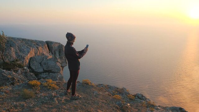 Circular shot from the back of a girl standing on the edge of a cliff who takes video and pictures with smartphone of beautiful smoke and a couple of clouds against the backdrop of sunset and sea.