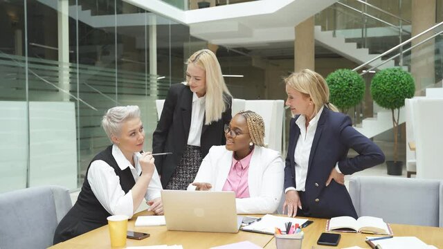 Three caucasian women and one african female manager coworking in light modern office, coworking, chatting and talking, using laptop.
