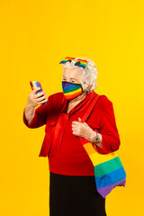 Studio portrait of a senior woman wearing a red shirt, rainbow sunglasses, rainbow colored facial mask and a bag, having a video call with her mobile phone, against a yellow background