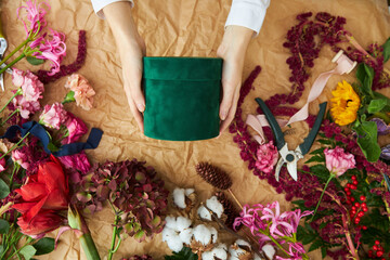 A female florist holds a dark green gift box. The surrounding frame is the colors and things of the florist. Craft paper parchment in the background.