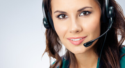 Portrait of smiling beautiful female phone operator in headset, green confident clothing, over grey...
