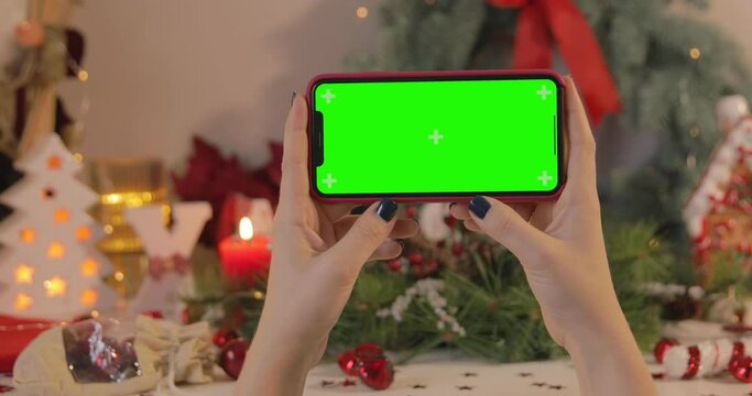 A woman holding an iPhone 11 Pro Max with a green screen on a Christmas background. Horizontal orientation. POV. Keyed Chroma key.