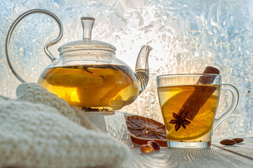 Winter warming tea with lemon and orange against the background of frosty patterns. Hot drink in a glass kettle and a cup on a wooden table in the background of the window.