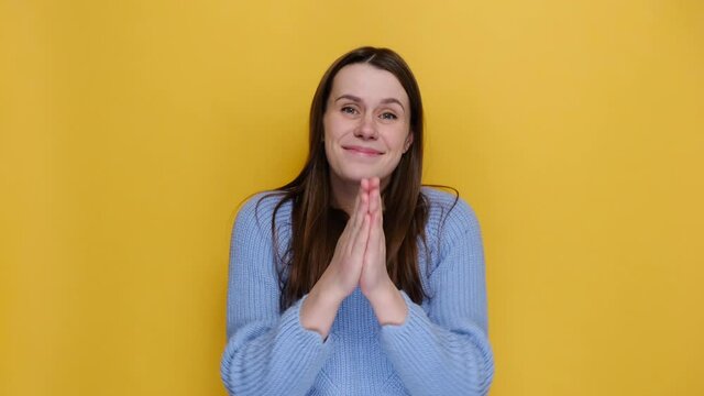 Pleading young woman hold hands folded in prayer wait for special moment keep fingers crossed making wish, dressed in blue sweater, isolated on yellow studio background. People lifestyle concept. 