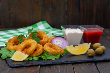 Deep fried squid or fish rings appetizer. Breaded squid or fish rings with lemon,  ketchup and mayonnaise. Golden, crunchy calamari snack on black board.