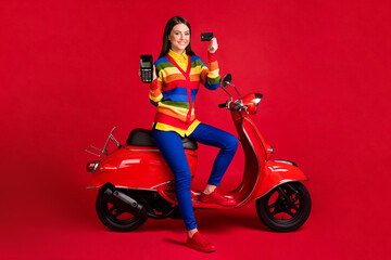 Plakat Photo portrait of happy woman holding credit card terminal in hands sitting on scooter isolated on vivid red colored background