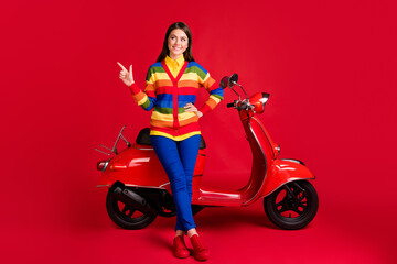 Plakat Photo portrait of dreamy girl pointing finger looking to side sitting on retro scooter isolated on vivid red colored background