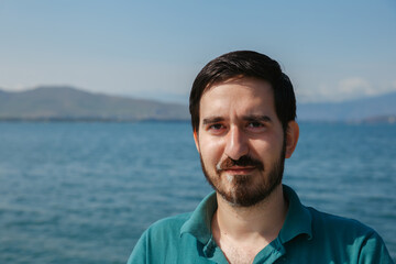 Young bearded man with charming smile standing on background of sea and enjoying summer vacation while looking at camera 