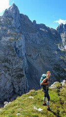 Fototapeta premium A woman standing at the edge of a mountain with a view on high mountain behind her. There are sharp mountains and high peaks around. The Alpine meadow is overgrown with lush green grass. Adventure