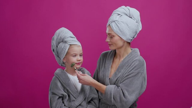 Mom and daughter doing skincare routine together in the bathroom. Mother massaging face of happy pleased kid relaxing looking funny. Purple background. Beauty concept.