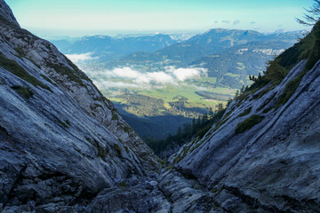 A panoramic view on from an Alpine top on a vast valley. There are sharp mountains and high peaks around. The Alpine slopes are almost barren. Lush green valley. Bright day. Serenity and freedom.