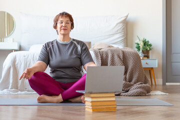 Senior woman meditates at home in a lotus position in front of a laptop monitor. The concept of a healthy lifestyle in old age.