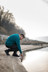 A young man in a turquoise sweater wets his hands in the river