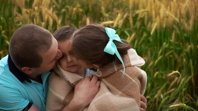 Portrait of father with son and daughter wrapped in blanket who sit in summer wheat field in slowmo. A man kisses and hugs his children. They are smiling and laughing. Concept of happy family outdoors