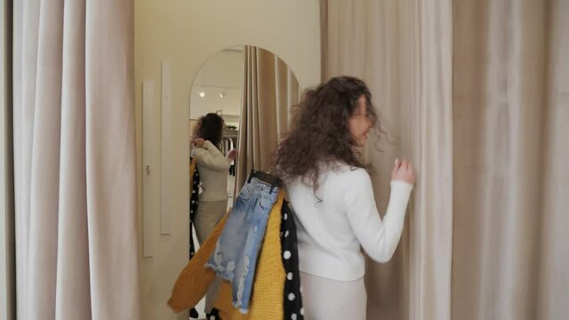 A beautiful girl with curly hair and a white sweater enters the fitting room holding hangers of clothes and closes the curtain she decides to try it on shopping concept.