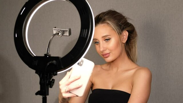 Beautiful woman takes a selfie on her phone at ring lamp in studio. Modern girl taking photos makeup for social networks.