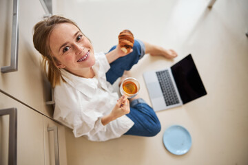 Joyful young woman having breakfast and using notebook at home