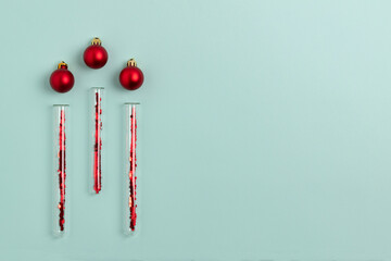 Medical test tubes with red sparkles inside lie next to Christmas balls on a mint green background. Festive flat lay with copy space.