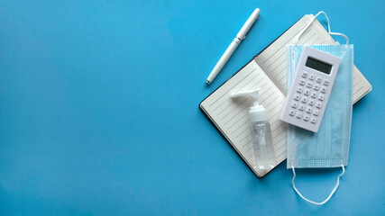 Top view of calculator,pen, notebook, face mask and a bottle of hand sanitizer isolated on a blue background with copy space.