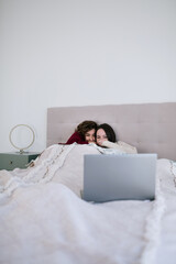 Two white teenage girls with brown hair in bed watching a movie on the computer. They are wrapped in a beige blanket