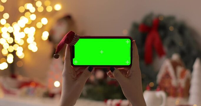 Girl Hands holding the iPhone Xr smartphone in horizontal orientation with green screen and Santa Claus' hat on top on Christmas background