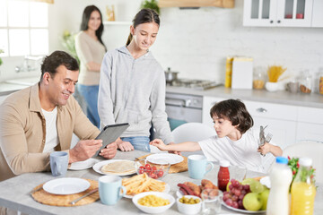 Cook to serve love. Middle aged latin husband using tablet pc, watching news, waiting for dinner while his children and wife serving table in the kitchen at home