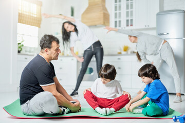 Fitness routine. Full length shot of two little boys sitting on a mat with father while their mom and sister exercising in the background