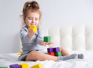 Little girl playing with colorful wooden cubes sitting on bed. Home activities for kids.
