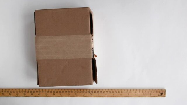 Taping delivery box with clear tape while preparing it for shipping. Wooden yardstick placed on the white surface next to the parcel