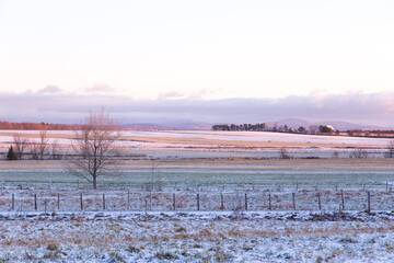 Fields covered in dusting of fresh snow, with a scattering of houses and mountains in soft focus background seen during a winter golden hour dawn, Quebec City, Quebec, Canada