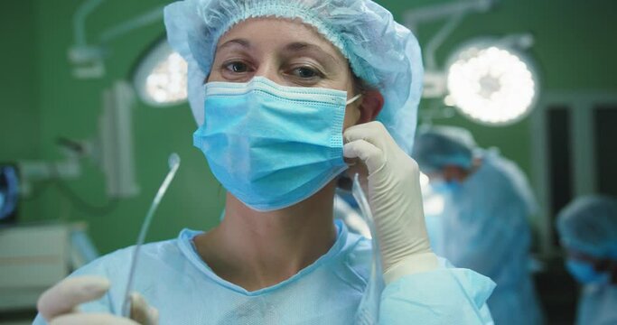 Close up portrait. Caucasian young female professional surgeon in medical uniform putting off mask, looking at camera and smiling. Team of surgeons performing operation in surgery room on background