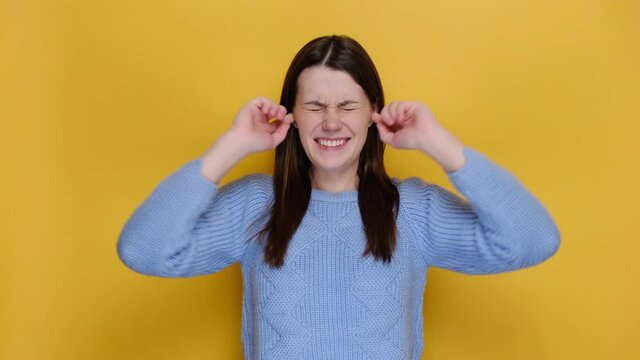 Crazy frustrated young caucasian woman cover ears with fingers keeping eyes closed screaming, dressed in blue sweater, posing isolated on yellow studio background. Emotion people lifestyle concept