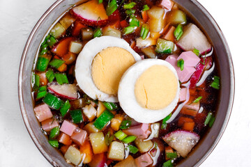 Russian cold soup Okroshka with vegetables, sausage, eggs and kvass in a bowl on the white table