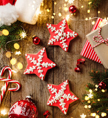 Christmas gingerbread stars, fir branches, red decorations, lights garland, Santa hat, orange and gift boxes on wooden background. Merry Christmas and New Year holiday. Xmas cookies, top view, bokeh