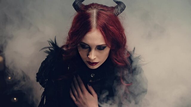Scary demon doing witchcraft. Red-haired girl witch with black horns sitting on floor in candlelight and praying with folded hands. Halloween mystery rite. Ritual of Occultism or esoterics concept.