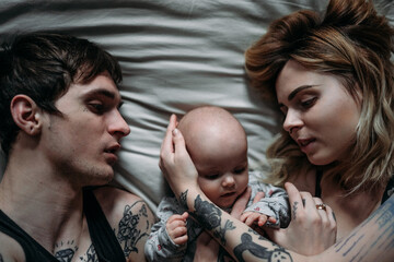 parents with a tattoo lie on the bed with a child and hug the face of a newborn with their hands...