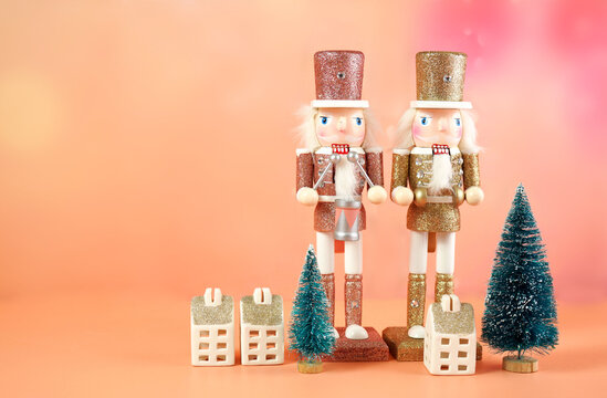 Bright modern Christmas scene with pink and gold wooden nutcracker ornaments, gifts and mini Christmas trees against a pastel coral background. Negative copy space.