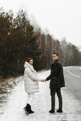 Fototapeta na wymiar Young couple holding each other hands in the background of the road and winter forest. Love, relationship, winter holidays. Winter couple photo ideas