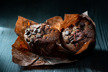 on a dark wooden background, with side grazing light, muffins in the foreground with pieces of dark chocolate. Still life