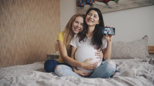 Portrait of two women showing an ultrasound image of future baby on smartphone and smiling to camera. Homosexual pregnant lesbian couple. IVF, LGBT Pride Month, relationship, childbirth, concept.