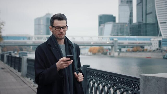 Man in black coat and eyeglasses with phone walking on background of business skyscrapers in Moscow, slow motion. Middle shot of young man in the city business area
