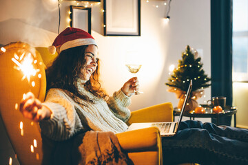 Happy young woman wearing Santa hat, drinking champagne, talking with friends on video call celebrate New Year party. - obrazy, fototapety, plakaty